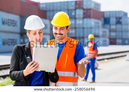 Manager with clipboard full of freight documents talking with worker on shipment yard in front of container