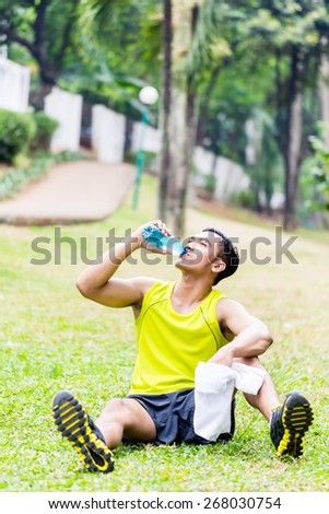 Asian man having break from sport training in tropical park, drinking water from a bottle
