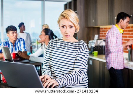 Chinese woman programmer in lounge of IT start-up with laptop, co-workers in the background