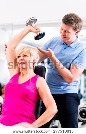 Senior woman at sport exercise with dumbbell in gym with trainer to gain strength and fitness