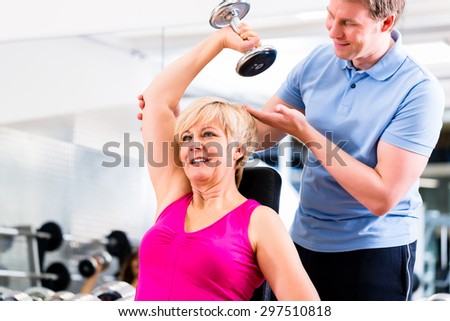 Senior woman at sport exercise with dumbbell in gym with trainer to gain strength and fitness