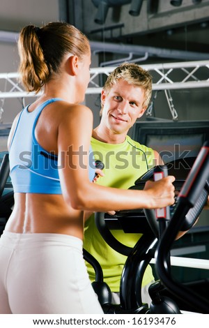 Couple in a gym, she is working out on a cross trainer, he is looking at her and could be her personal trainer supervising her training