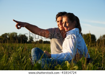 Young couple sitting together on a green meadow in summer looking into a golden sunset contemplating and daydreaming their future (marriage, children, divorce, eventual death and so on)