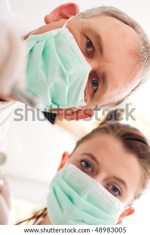 Dental treatment with dentist and dental assistant bowing over the patient as seen from patient\'s perspective. They have drills and angled mirrors, focus on eyes of dentist