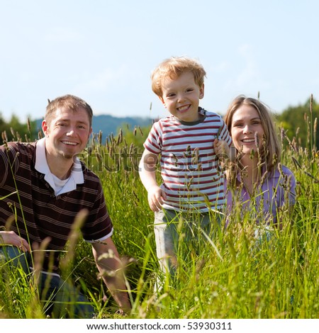 Very happy family with son sitting in a  meadow in the summer sun in front of a forest and hills, they are nearly hidden by the high grass, very peaceful scene