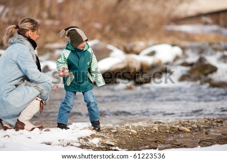Family - mother and son to be seen - on a walk along a riverbank in winter; the child is throwing a snowball