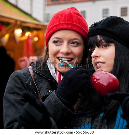 Two women on Christmas market eating apple and candy in front of a booth, it is cold
