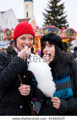 Two women on Christmas market eating cotton candy in front of a booth, it is cold
