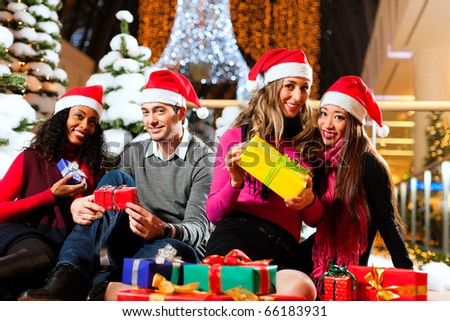 Group of four people - diversity - with Santa hats sitting amid artificial snow covered fir trees and lights with Christmas presents in a shopping mall