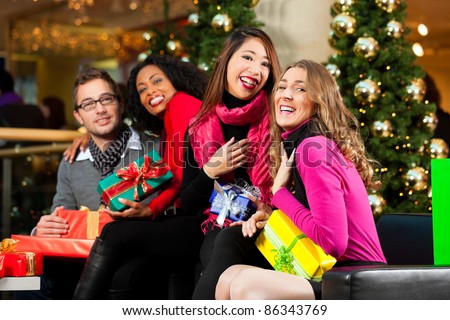Diversity group of four people - Caucasian, black and Asian - sitting with Christmas presents and bags in a shopping mall in front of a Christmas tree with baubles