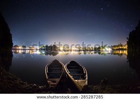 Two boats at the shore of a serene lake at night. There are lights of the city on the background, behind the Phewa lake - the most famous lake in Pokhara (Nepal).