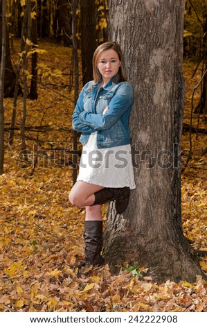 full length portrait of young female teen leaning against tree arms crossed in forest during autumn