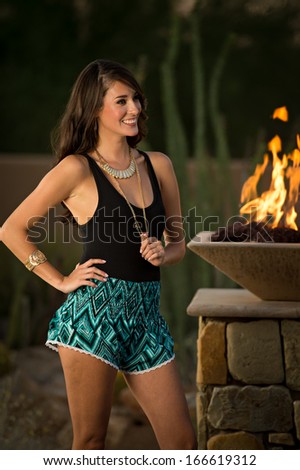 Beautiful brunette fashion model standing next to a stone column with a fire feature on top posing outdoors.