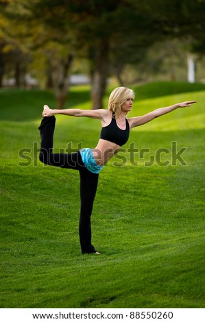 Beautiful blond young woman practicing a yoga pose for balance on one leg in the park on a green lawn with trees in the distance.