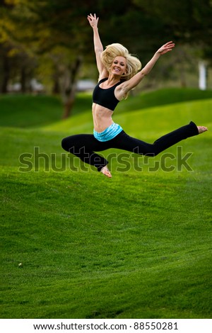 Beautiful blond young woman practicing yoga in the park on a green lawn with trees in the distance.  She is jumping in the air with her arms overhead.