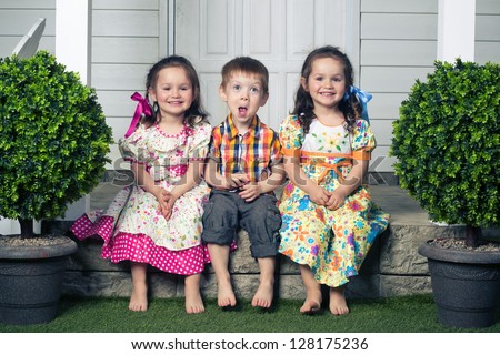 two beautiful twin girls and one red head boy are sitting together on the porch of white mansion house