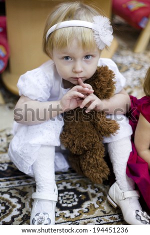little kinder garden girl in white dress sitting on the carpet with toy bear