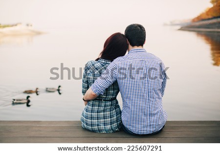 Rear view of a romantic young couple sitting on the river dock in autumn