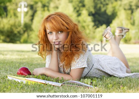 Redhead woman lies on green grass and reads green  book, covers the face during spring / summer time . Happy smiling beautiful young university student studying lying down in grass with red apple.