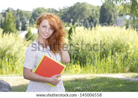 Redhead woman standing in park near university, college with books on nature in summer or spring. Happy smiling beautiful young girl university student