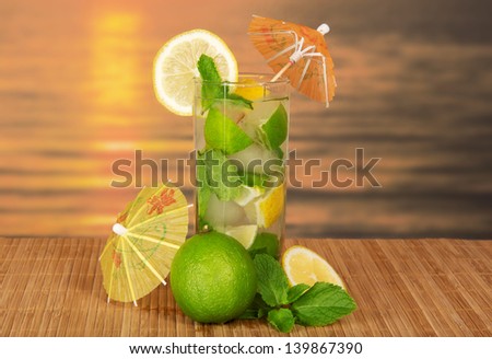 Green cocktail of alcohol with an umbrella, a lemon and spearmint, on a bamboo cloth against the sunset