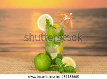 The glass of a cold mojito, is decorated with an umbrella, the spearmint, on a bamboo cloth against the sunset