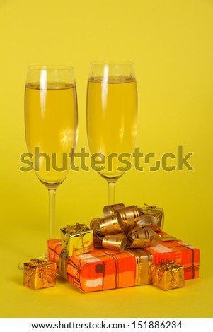 Wine glasses with champagne, a gift box with a beautiful bow and souvenirs on a yellow background