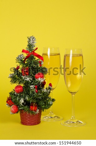 Small Christmas fir-tree in a pot, and two wine glasses with champagne on a yellow background