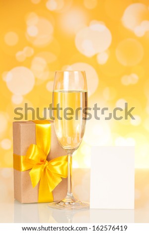 Glass with champagne, a gift and an empty card on an abstract yellow background