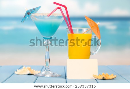 Glass of juice, cocktail and blank card on sea background