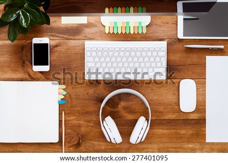 Brown wooden table, top view. On the table are, stationery items, notebook, gadgets, green flower, stickers, keyboard, mouse, white paper, headphones for convenient operation.