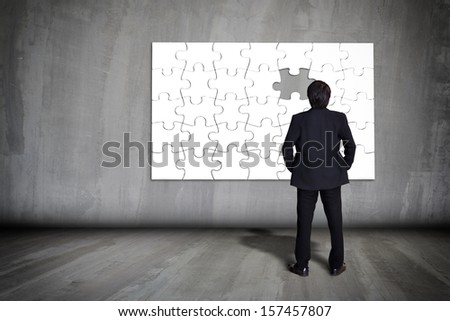 Businessman figuring out puzzle pieces with piece missing on grey wall