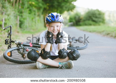 Happy girl with bike, helmet and protection sitting on the road at the park, summertime