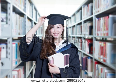 asian female student holding book and wearing academic dress in library