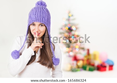 Cute young Caucasian woman with purple beanie hat smiling gesturing hush looking at camera with colorful decorated Christmas tree and presents blurred in the background.