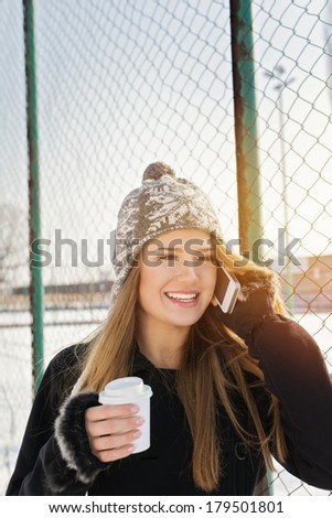 Cute young Caucasian teenage woman talking on the phone laughing holding a takeaway coffee cup smiling. Outdoors winter portrait of beautiful teenage girl speaking on the phone.