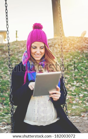 Happy young Caucasian pregnant woman with tablet computer outdoors sitting on swing in park on a sunny winter day. Mother expecting a baby using digital tablet enjoying outdoor.
