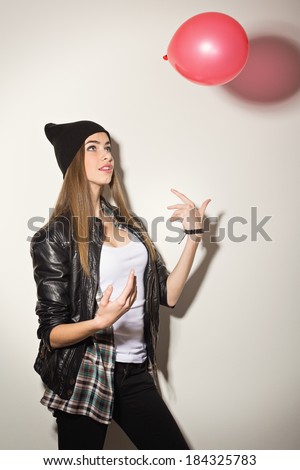 Cute hipster teenage girl wearing black jacket and beanie hat looking up throwing red balloon playing and having fun.