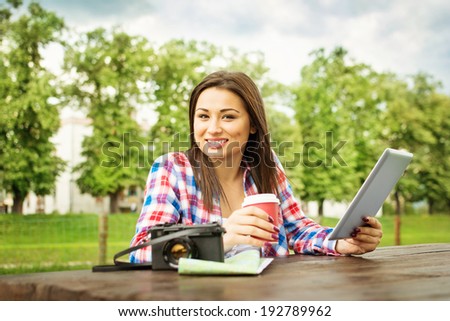 Young mixed race teenage tourist woman in park on summer day with tablet, map and camera drinking coffee. Traveling concept.