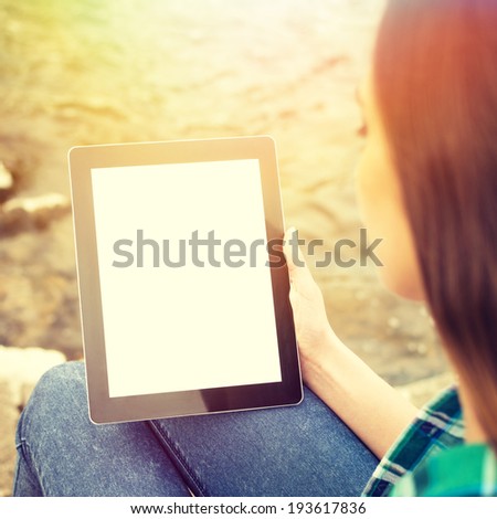 Young Caucasian brunette woman with digital tablet with isolated blank screen sitting by the river in summer.Square image with instant looking filter.