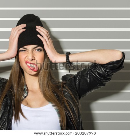 Hipster teenage girl making faces and holding her head wearing black beanie hat, leather jacket and bracelet against gray stripes background.