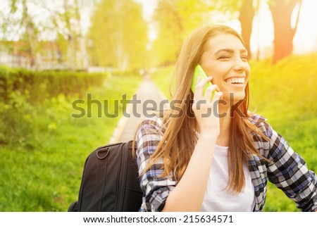 Happy teenage girl talking on the phone outdoors laughing. Cute young female student speaking on the phone in park on sunny summer day.