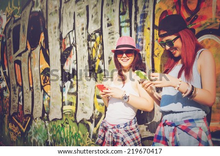 Closeup of two teenage girls with smart phones. Two young women using smartphones against colorful graffiti wall.