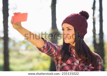 Young Caucasian brunette woman with knitted maroon colored beanie hat taking a selfie with smart phone in park. No retouch.