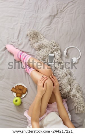 Woman in cozy pajama shorts and pink socks relaxing at home in bed. Top view of young woman\'s legs on gray sheet with smartphone, headphones, cookies and apple.