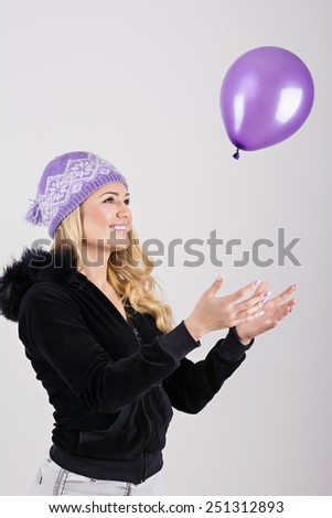 Happy Caucasian blonde teenage girl in purple knit beanie hat playing with purple balloon. Studio portrait, closeup, solid gray background, mild retouch, vertical.