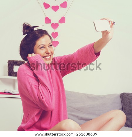 Beautiful young brunette woman with messy bun in pink sweater sitting in bed smiling taking a selfie. Young woman taking a self portrait with smartphone at home. Square format, instagram filter look.