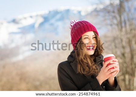 Happy young Caucasian brunette woman with red lips and red beanie hat with takeaway coffee outdoors in winter. Snowy mountain blurred in the background. Retouched, natural light, copy space.