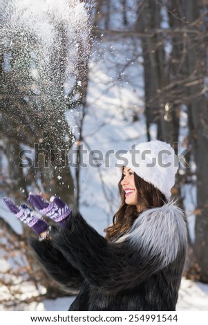 Happy young woman playing with snow outdoors on sunny winter day. Fashionable girl with fuzzy hat and coat throwing snow playing having fun. Vertical, no retouch, copy space.