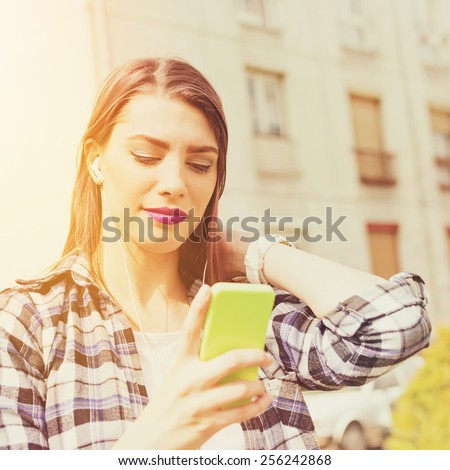 Closeup outdoors portrait of beautiful millennial teenage girl with smartphone texting outdoors in summer. Young woman with cellphone and earphones, urban background, filter, retouched, square format.